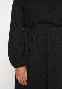 Svart klänning med texturerad tyg som har prickar, en åtsittande midja med bälte, och långa puffärmar. Enkla knappdetaljer.