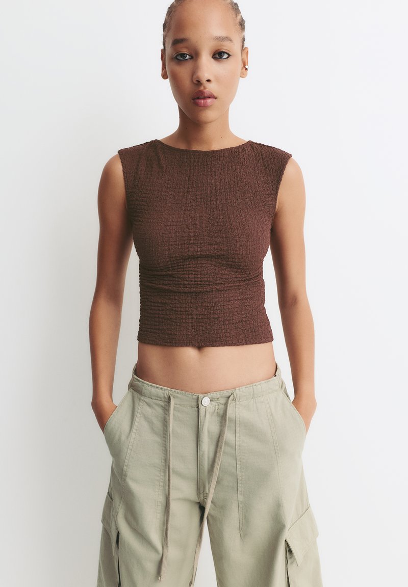 Top cropped de culoare maronie, ajustat, dintr-un material texturat. Asortat cu pantaloni cargo lejeri, de culoare verde deschis, cu buzunare laterale și talie cu șnur.