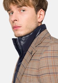 Brązowa marynarka w kratę z pomarańczowymi i niebieskimi akcentami, nałożona na granatowy kołnierz puchowy, prezentująca teksturowane materiały i elegancki design.