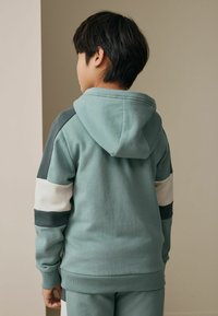 Hanorac cu glugă în nuanță teal estompată, cu dungi contrastante în teal închis și alb pe mâneci; materialul este moale și texturat, cu manșete ribbed.