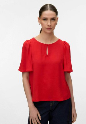 Kobieta o ciemnych włosach związanych nisko w kucyk, ubrana w czerwoną bluzkę z krótkim rękawem i dekoltem z wycięciem oraz ciemne spodnie, stojąca na jednolitym białym tle.