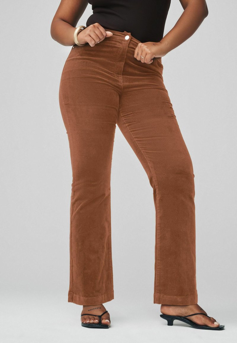 Braune Corduroy-Hosen mit Schlag und hohem Bund, vorderem Knopfverschluss und glatter Textur. Kombiniert mit schwarzen Sandalen.