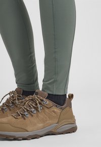 Bruine wandelschoenen van gestructureerd suède met grijze zolen, gecombineerd met strakke groene leggings met zijnaad en een gladde afwerking.