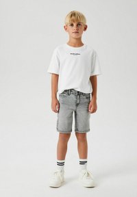 Vit kortärmad t-shirt med texten "Studio edition", kombinerad med ljusgrå denimshorts. Vita sneakers och svartrandiga strumpor fulländar looken.