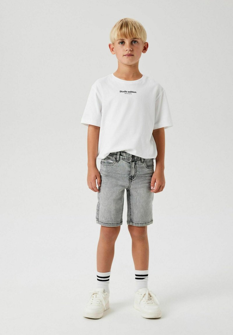 Vit kortärmad t-shirt med texten "Studio edition", kombinerad med ljusgrå denimshorts. Vita sneakers och svartrandiga strumpor fulländar looken.
