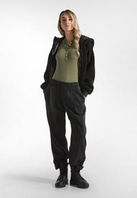 Jachetă neagră din fleece peste un henley verde cu mânecă lungă și butoane, asociată cu pantaloni negri largi și cizme negre cu șireturi, pe un fundal alb.