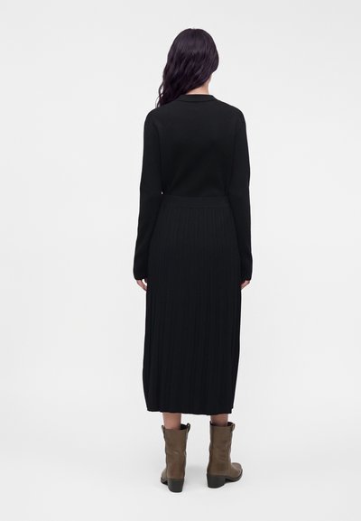 Femme aux longs cheveux noirs portant une robe noire à manches longues et des bottes marron mi-mollet, debout, dos à un fond uni.