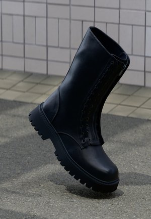 Schwarzer Leder-Kampfstiefel mit dicker Profilsohle und seitlichem Reißverschluss, aufrecht stehend auf gepflastertem Untergrund vor einer gefliesten Wand.