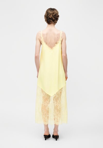 Άτομο που φαίνεται από πίσω, φορώντας ανοιχτό κίτρινο αμάνικο φόρεμα με δαντελένιες λεπτομέρειες και μαύρα σανδάλια με τακούνι, στέκεται σε απλό φόντο.