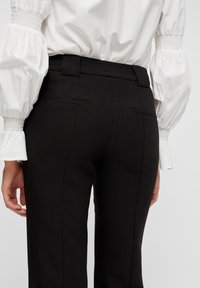 Svarta skräddarsydda byxor med smal passform, med bakfickor, en slät yta och midjeband. Vit blus med puffärmar delvis synlig.