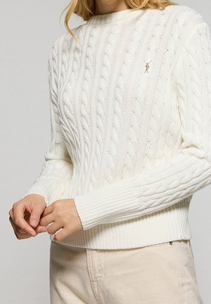 Femme portant un pull en maille torsadée blanc avec un petit logo brodé, associée à un pantalon beige clair, vue du cou aux hanches.