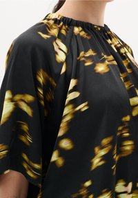 Blouse noire avec un motif floral abstrait dans des teintes dorées et roses. Présente un col froncé et des manches amples et fluides. Texture lisse.