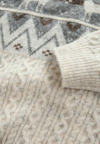 Strickpullover in Creme mit einem strukturierten Kabelmuster, ausgestattet mit einem grau-braunen Zickzackdesign auf den Schultern und gerippten Bündchen.