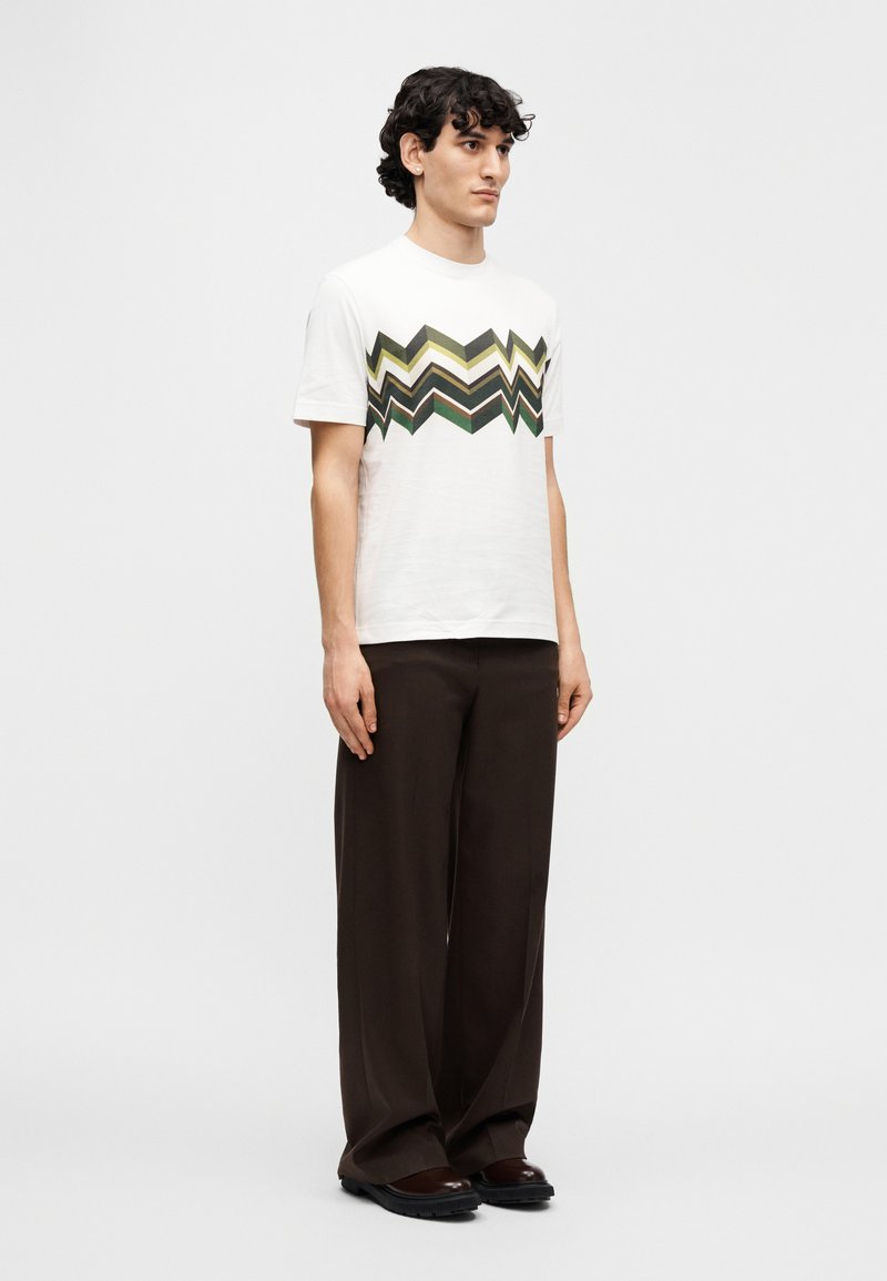 White cotton t-shirt with zigzag pattern in olive green, yellow, and brown; paired with loose-fitting brown trousers and dark shoes.