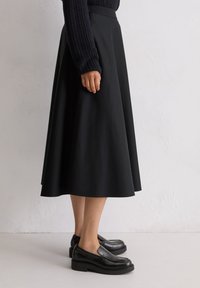 Jupe noire évasée, longueur genou, en tissu lisse. Associée à des chaussures noires à enfiler, avec une plateforme basse et un bout rond.