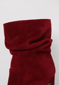 Botín de ante rojo hasta el tobillo con puño doblado y superficie texturizada. Presenta una punta redondeada y sutiles detalles de costura.