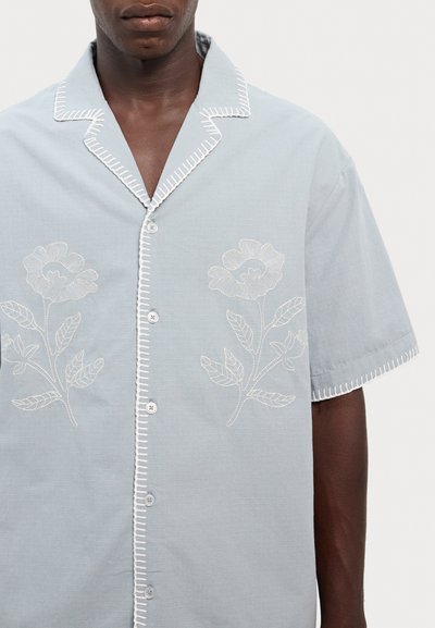 Šviesiai mėlynas trumpomis rankovėmis marškinėliai, puoštas baltais siuvinėtus gėlėmis ir lapų raštais, su iškirpte ir mygtukų uždarymu, papuoštu kontrastinga siūle.