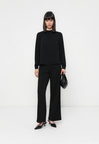 Black long-sleeve top with a relaxed fit, paired with black wide-leg trousers. Black pointed-toe heeled shoes and a small black handbag.