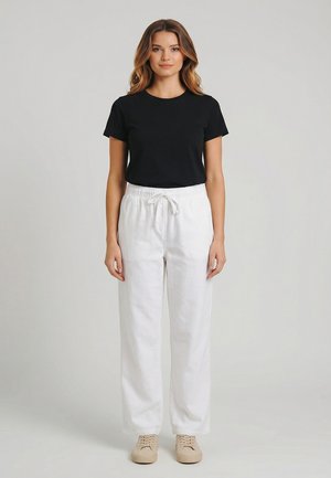 Donna in piedi di fronte, indossa una maglietta nera a maniche corte, pantaloni bianchi con coulisse e sneakers beige su uno sfondo semplice.