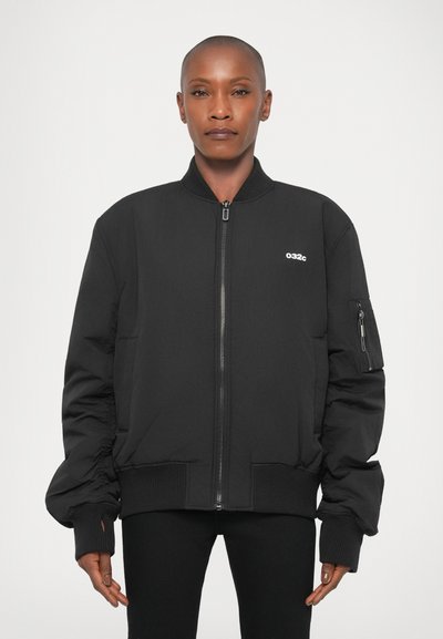 Person with shaven head wearing a black zip-up bomber jacket with a small white "032c" logo on the chest against a plain background.