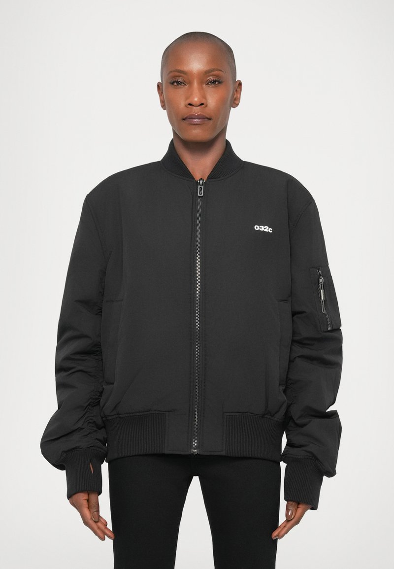 Osoba s oholenou hlavou, oblečená v černé bomber jacket na zip s malým bílým logem "032c" na prsou, na jednobarevném pozadí.