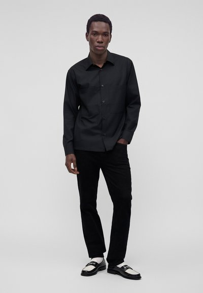 Homme portant une chemise noire boutonnée, un pantalon noir, des chaussettes blanches et des mocassins noirs et blancs, debout devant un fond clair uni.