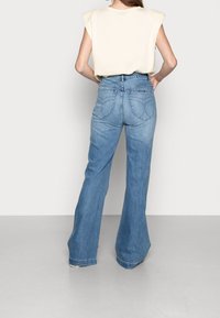 Jeans acampanados de color azul claro con cintura alta y detalles en los bolsillos traseros, combinados con una blusa sin mangas color crema. Textura suave de mezclilla.