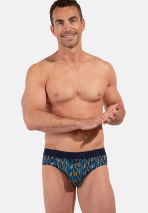 Lächelnder muskulöser Mann, der blaue gemusterte Slips mit schwarzem Bund trägt, vor einem einfachen hellen Hintergrund steht und in die Hände klatscht.