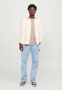 Lys beige skjorte med knapper, lys rosa t-skjorte, lyseblå jeans og marineblå og hvite joggesko. Håret er kort og krøllete.