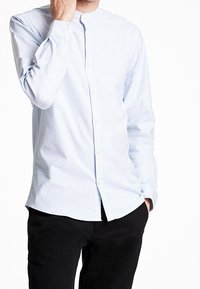 Camicia a maniche lunghe a righe blu chiaro e bianco con colletto a banda, chiusura a bottoni e silhouette aderente. Indossata con pantaloni neri.