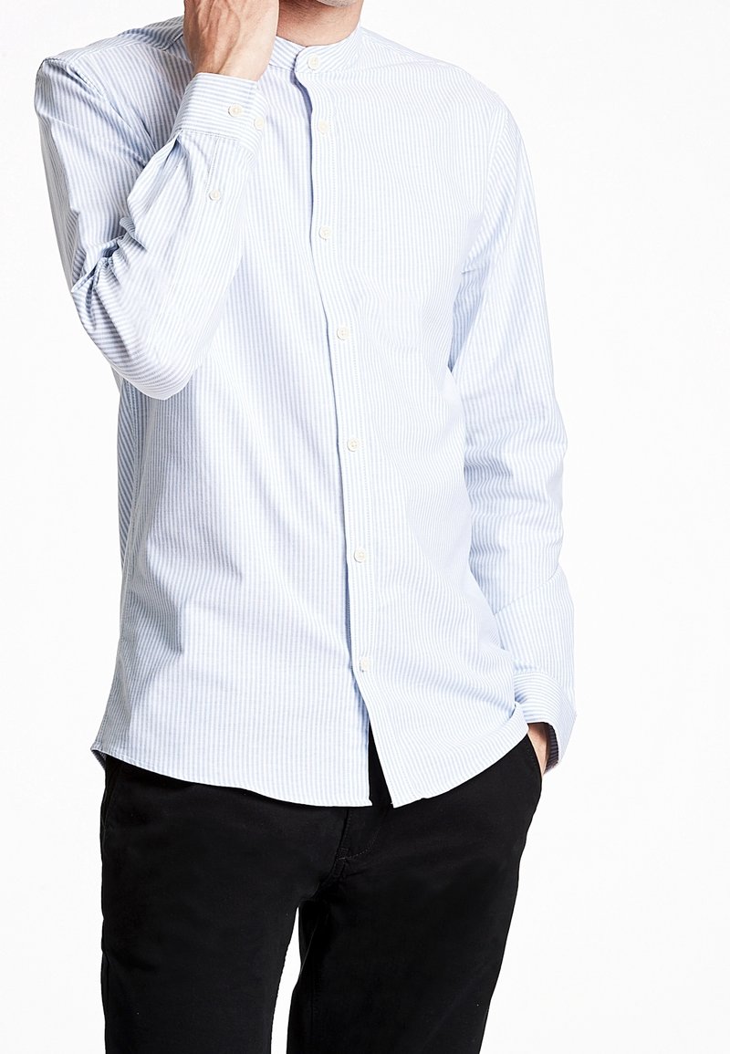 Camicia a maniche lunghe a righe blu chiaro e bianco con colletto a banda, chiusura a bottoni e silhouette aderente. Indossata con pantaloni neri.