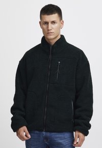 Black fleece jacket with a high collar, zippered chest pocket, and full front zipper. Features a textured, plush fabric and loose fit.