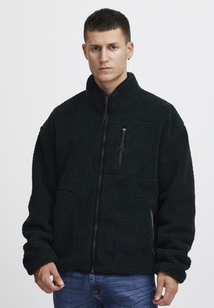 Black fleece jacket with a high collar, zippered chest pocket, and full front zipper. Features a textured, plush fabric and loose fit.