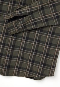 Camicia in flanella a quadri verdi con motivo a scacchi neri e beige. Presenta una chiusura a bottoni e maniche lunghe con polsini abbottonati.