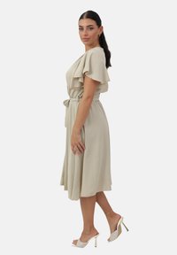 Vestido beige con mangas acampanadas, cintura entallada y dobladillo a la altura de la rodilla. Combinado con tacones blancos abiertos con un acento dorado.