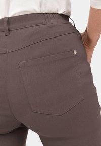 Pantalons gris-brun avec une taille élastique et une seule poche arrière. Tissu lisse avec une couture ton sur ton et un détail de bouton argent.