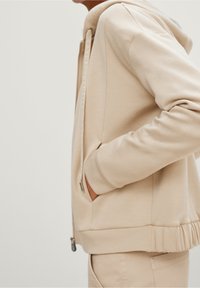 Beige hoodie van zacht materiaal, met een rits sluiting, zijzakken en een geribbelde zoom. De trekkoorden tonen de branding "CASUALIDENTITY".