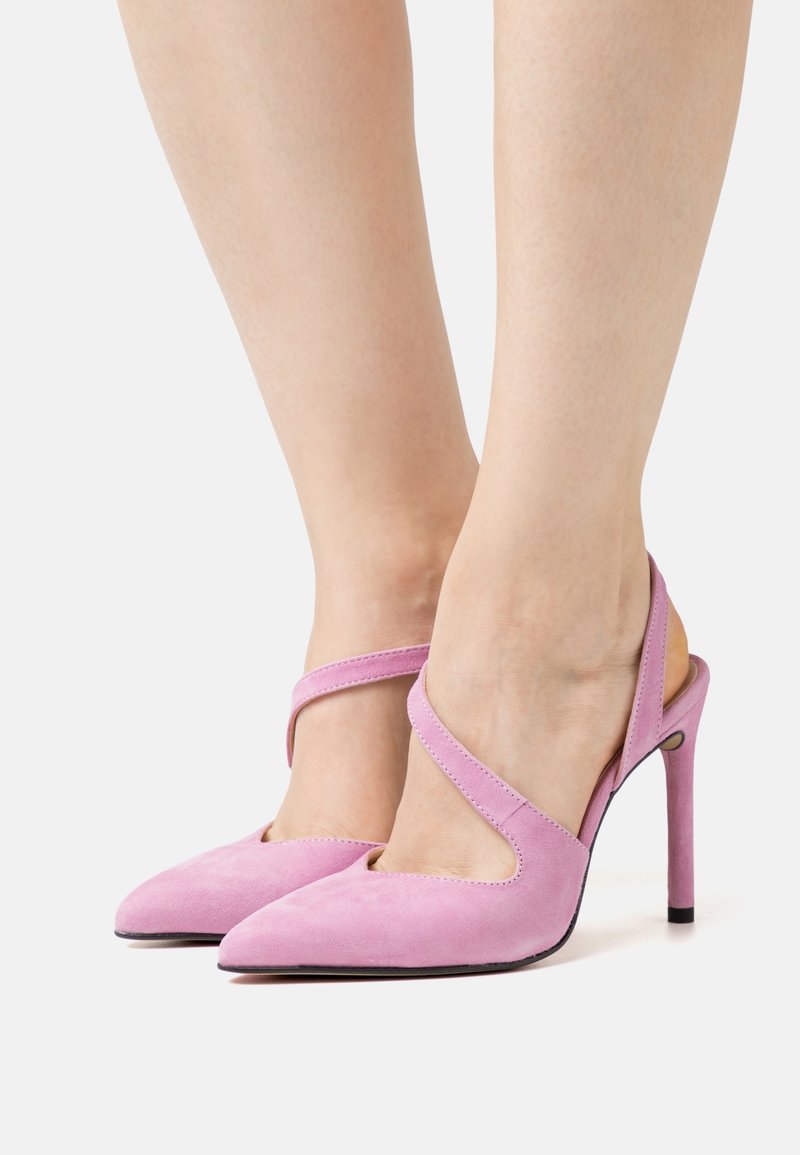 Pieds portant des chaussures à talons hauts en suède rose avec des bouts pointus et une bride unique traversant le dessus de chaque pied.
