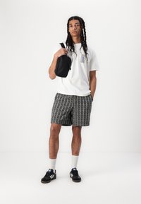T-shirt blanc avec une étiquette graphique, shorts noirs à motifs avec des rayures verticales, sac noir, baskets noires et chaussettes blanches.