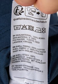 Hoito-ohjeet, joissa on tekstiilitietoja useilla kielillä ja pesuohjeita ilmentäviä symboleja. Teksti on pääasiassa valkoista kangasta vasten.