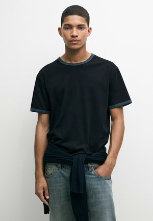 Junger Mann mit kurzen dunklen Haaren, der ein schwarzes T-Shirt mit dunklem Rand und einen dunklen Pullover um die Taille herum über blauen Jeans trägt.