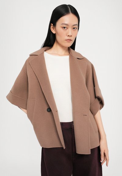 Veste oversize en mélange de laine marron avec un col cranté, fermeture à un bouton et poches avant, associée à un haut blanc et un pantalon sombre.