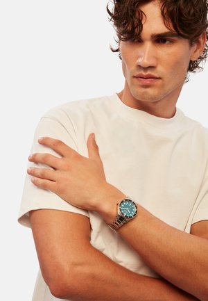 Sølv metal ur med en teal skive og lysende markeringer, båret på et håndled, parret med en simpel hvid t-shirt. Hudfarve lys beige.