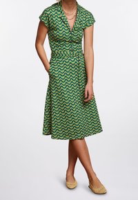 Grönmönstrad klänning med korta ärmar, V-ringning och figurnära midja. Anmärkningsvärd vågmönstring i gult och blått. Ett par vävda neutrala ballerinas.