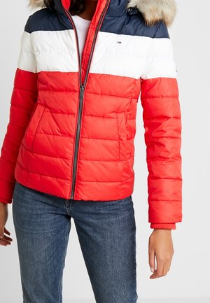 Mujer con una chaqueta acolchada en rojo, blanco y azul marino con capucha forrada de piel y vaqueros azules, posando contra un fondo blanco.