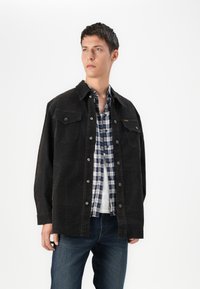 Chaqueta de mezclilla negra con dos bolsillos en el pecho, llevada sobre una camisa de cuadros en azul y blanco. El modelo también usa jeans azules, con una capa interior blanca lisa.