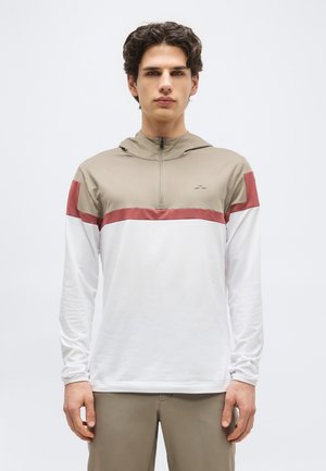 Νέος άνδρας με σκούρα, σγουρά μαλλιά, φορώντας ένα μπεζ, σκουρόχρωμο κόκκινο και λευκό μακρυμάνικο πλεχτό με φερμουάρ στο ένα τέταρτο και μπεζ παντελόνι, στέκεται μπροστά σε απλό φόντο.