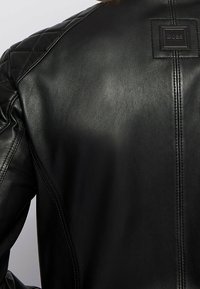 Černá kožená bunda s prošívanými detaily na ramenou, tónovaným šitím a čtvercovou nášivkou s logem poblíž výstřihu. Celkově hladká textura.