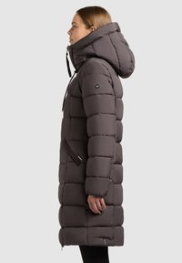 Lang, fyldig, mørkegrå parka med hætte. Har en quiltet design, frontlynlås og elastiske snore til justering.