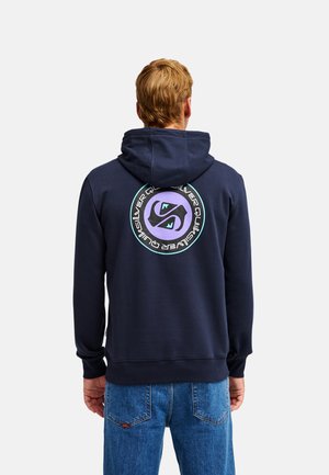 Mann mit kurzen blonden Haaren, der einen dunkelblauen Hoodie mit einem runden lila-schwarzen Motiv auf dem Rücken und eine blaue Jeans trägt.